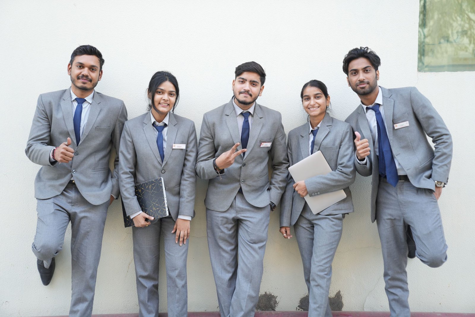 Group of young professionals in formal suits posing with smiles and confidence against a wall.